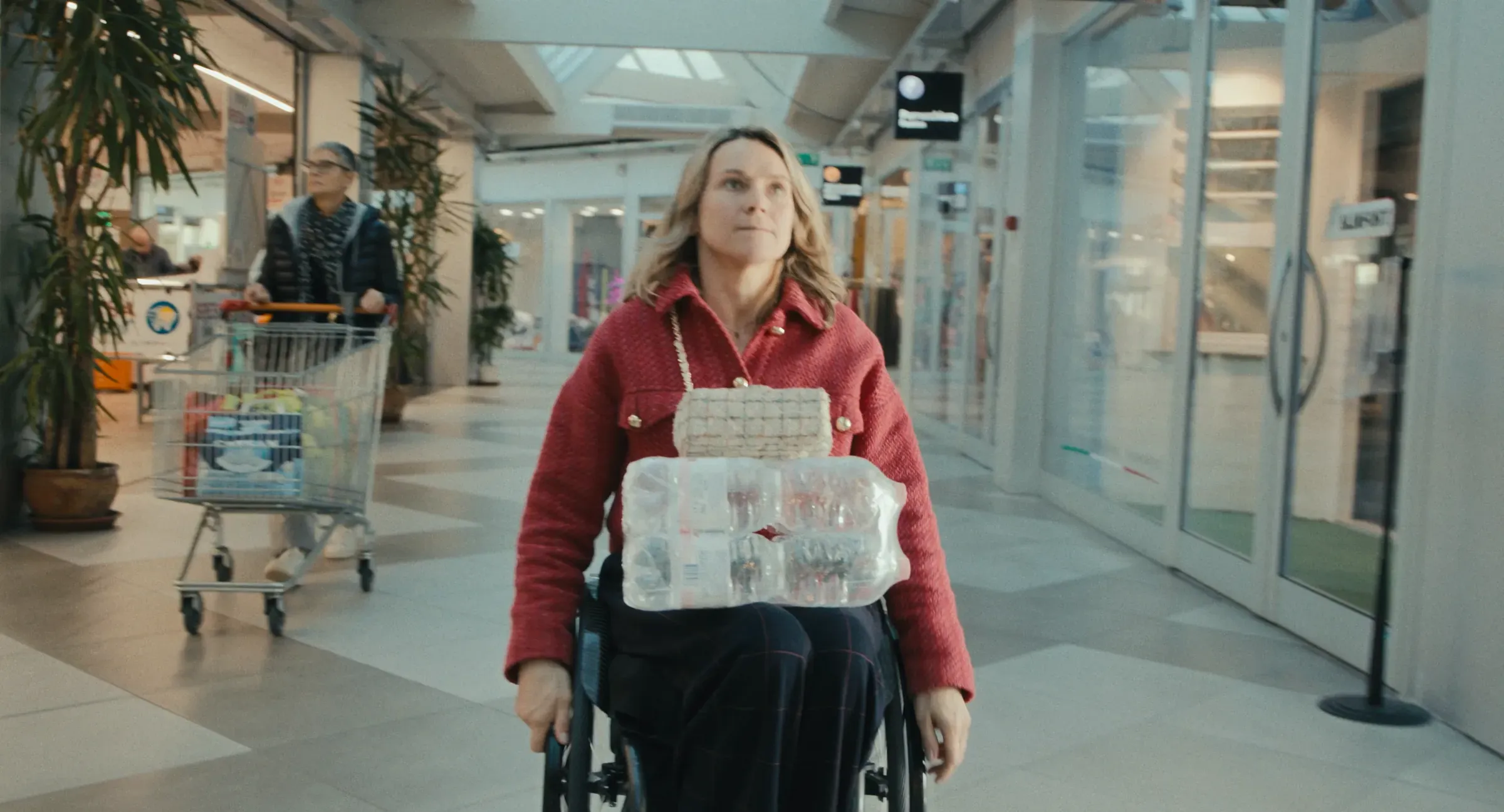 Film still from Breaking Barriers: Svetlana Moshkovich crossing a shopping arcade in her wheelchair, a pack of water bottles on her lap